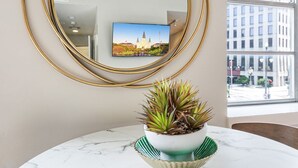 A room with a round mirror, a television displaying a cityscape, and a potted plant on a marble table.