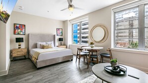 A modern bedroom with a bed, bedside tables, a round mirror, a dining table, and a view of buildings through large windows.
