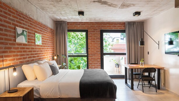 A modern room with a brick wall, a bed with white and gray bedding, a desk with a chair, and a large window with curtains. - H5063 (Buenos Aires)