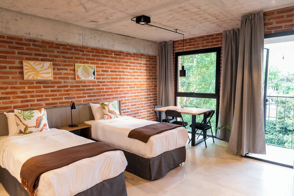 A modern hotel room with two beds, a small table, and a large window with curtains.
