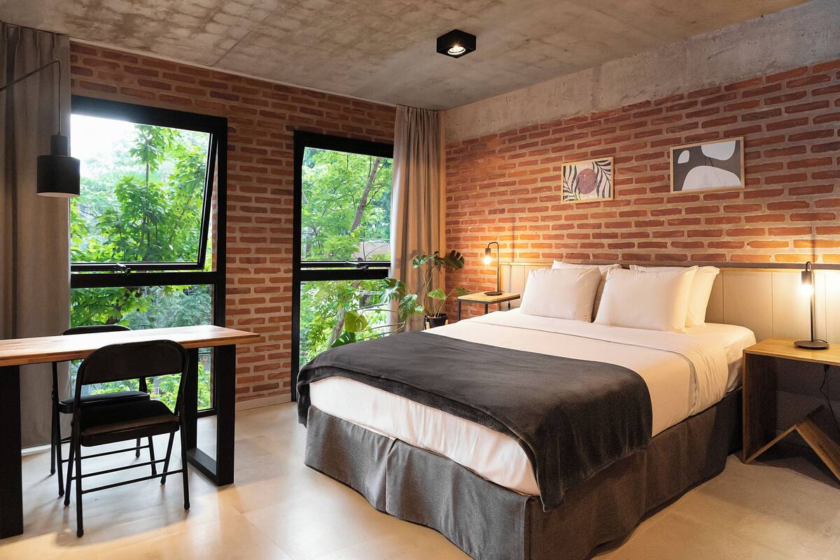 A bedroom with a brick wall, a desk, a chair, a bed with pillows, and a bedside table with a lamp.