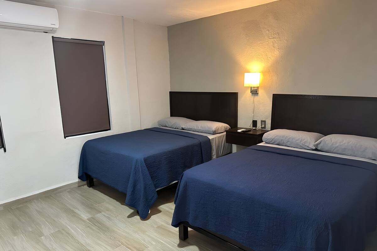 A hotel room with two beds, a bedside table, a lamp, and a window with blinds.
