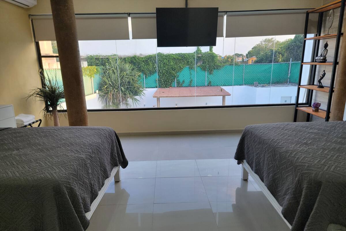 A hotel room with two beds, a large window, a TV, and a view of a green fence and buildings outside.