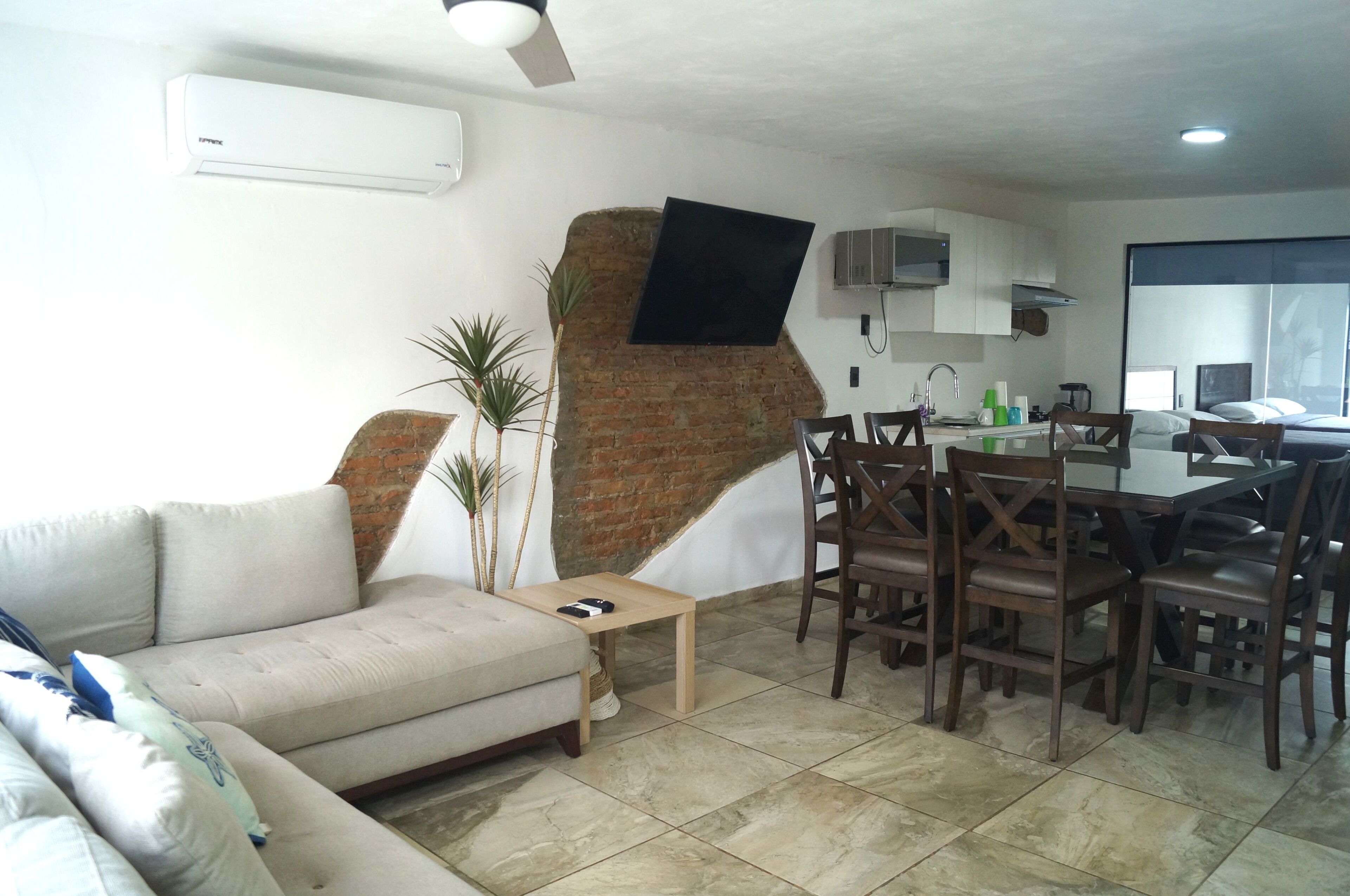 A living room with a sofa, a dining table with chairs, a TV mounted on a brick wall, and an air conditioning unit.