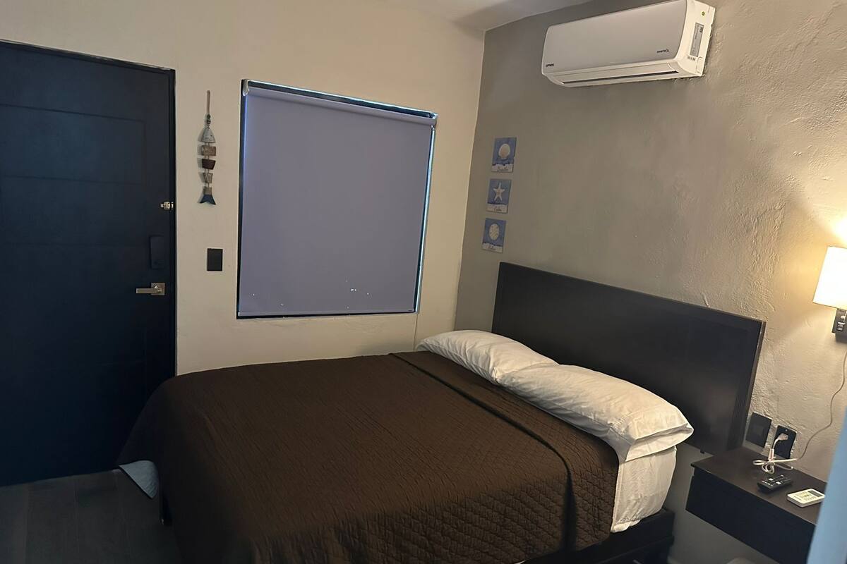 A hotel room with a bed, a bedside table with a lamp, an air conditioning unit, and a door.
