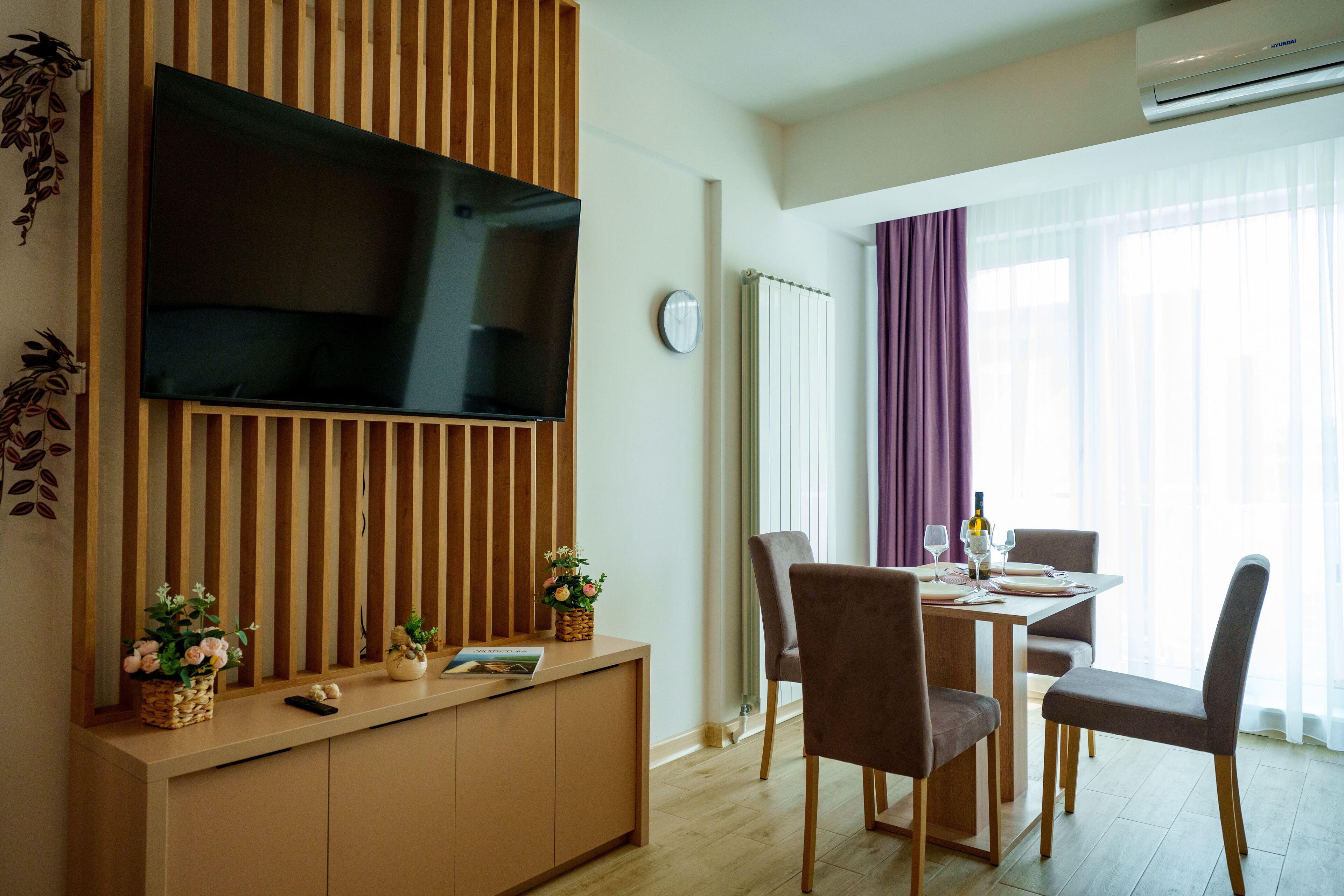 Un salon moderne avec un téléviseur à écran plat fixé sur un panneau en bois, une table à manger avec des chaises et une fenêtre avec des rideaux.