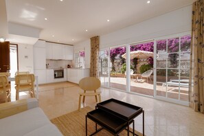 A spacious living area with a sofa, a coffee table, and a dining table with chairs. There is a kitchen area with white cabinets and a view of a garden through large glass doors. - Casa Andasol - holiday home close to the beach in Costabella (Marbella)