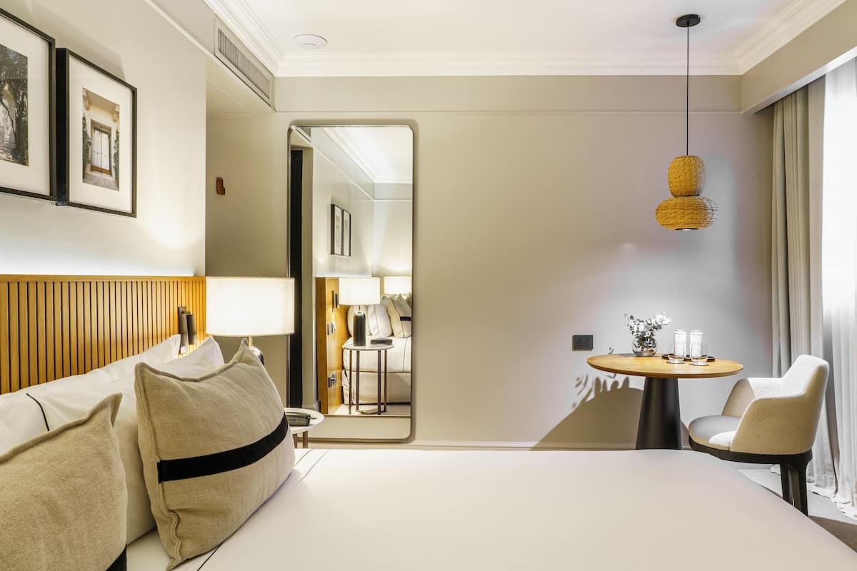 Habitación de hotel con cama, una silla, una mesa redonda con lámpara y un espejo que refleja otra habitación.