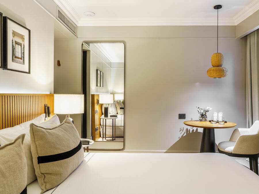 Habitación de hotel con cama, una silla, una mesa redonda con lámpara y un espejo que refleja otra habitación.