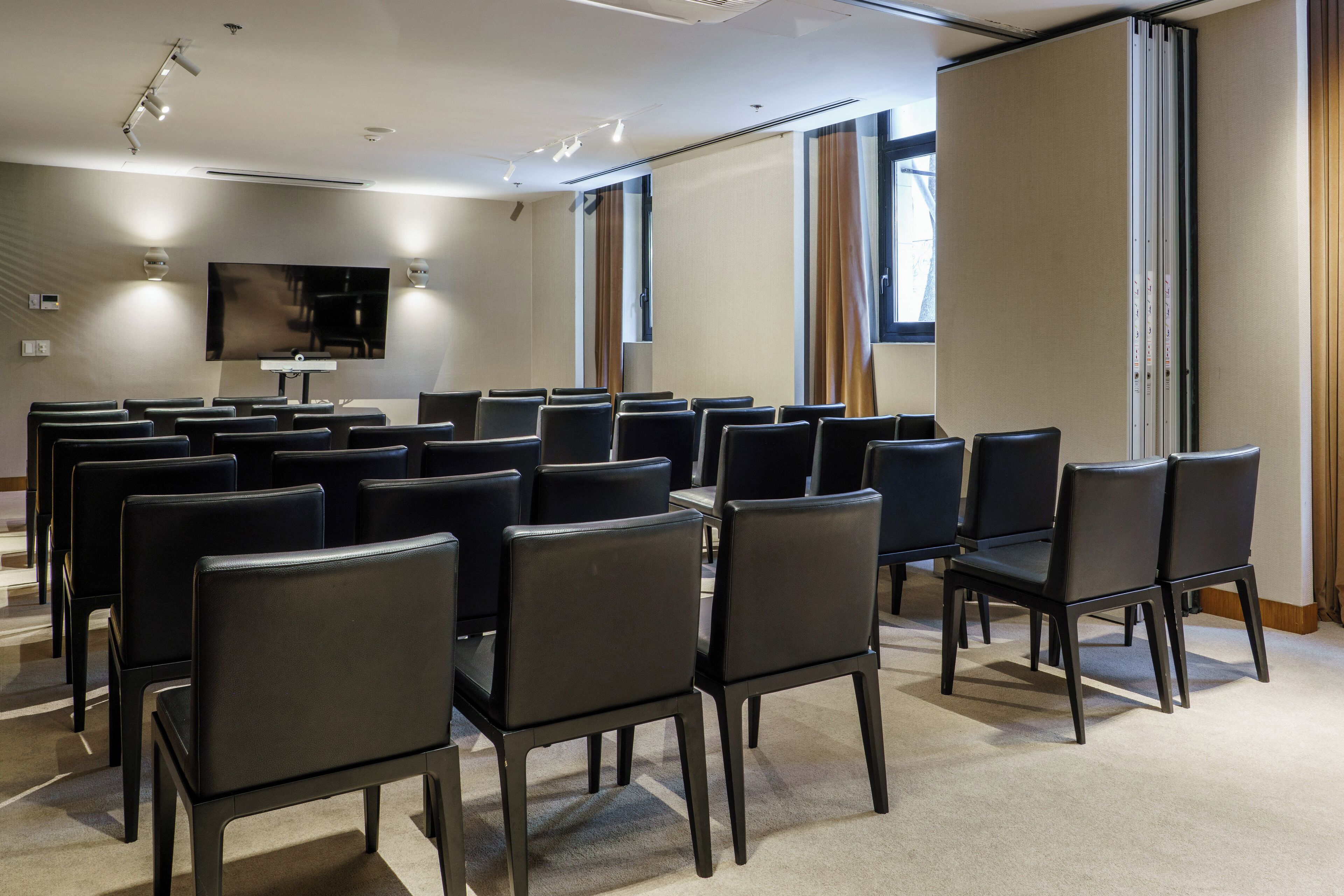 A conference room with rows of black chairs, a large flat-screen TV mounted on the wall, and a window with curtains.
