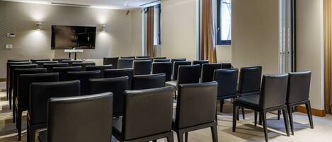 A conference room with rows of black chairs, a large flat-screen TV mounted on the wall, and a window with curtains.