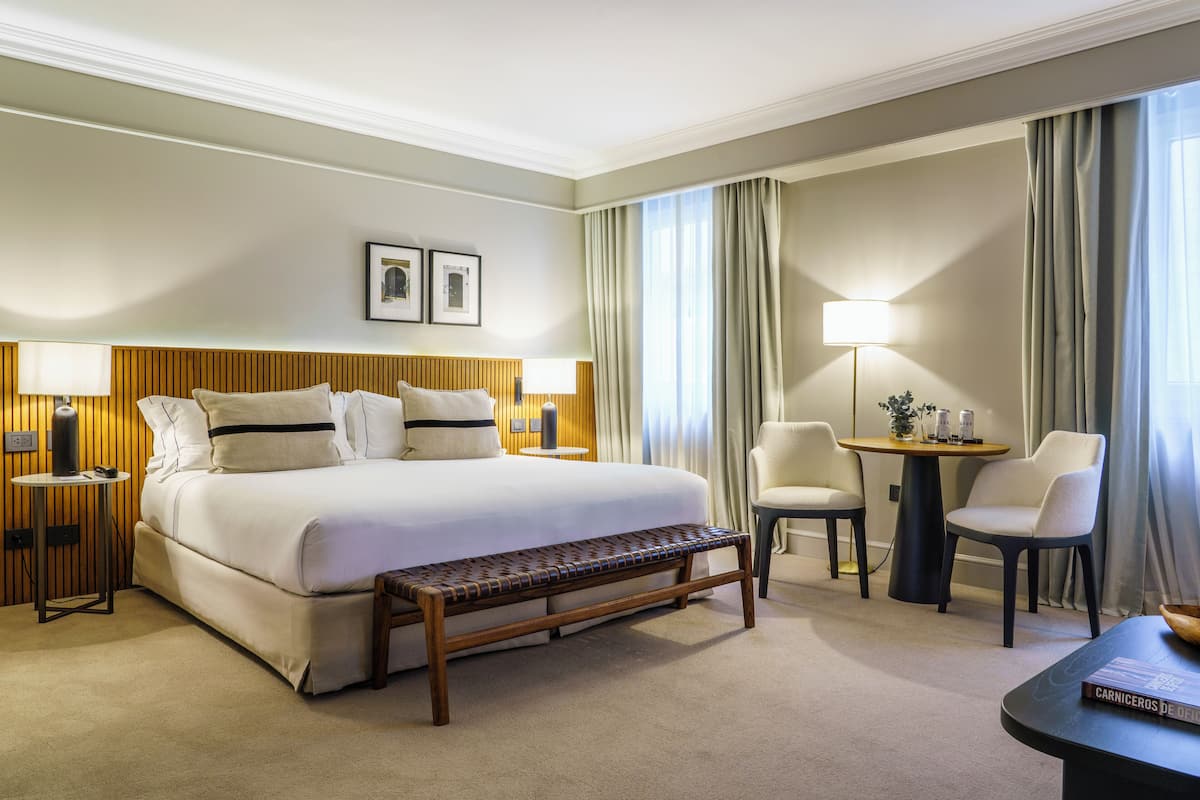 Habitación de hotel con una cama grande, mesitas de noche, un banco, una pequeña mesa con lámpara, dos sillas y vistas al exterior.