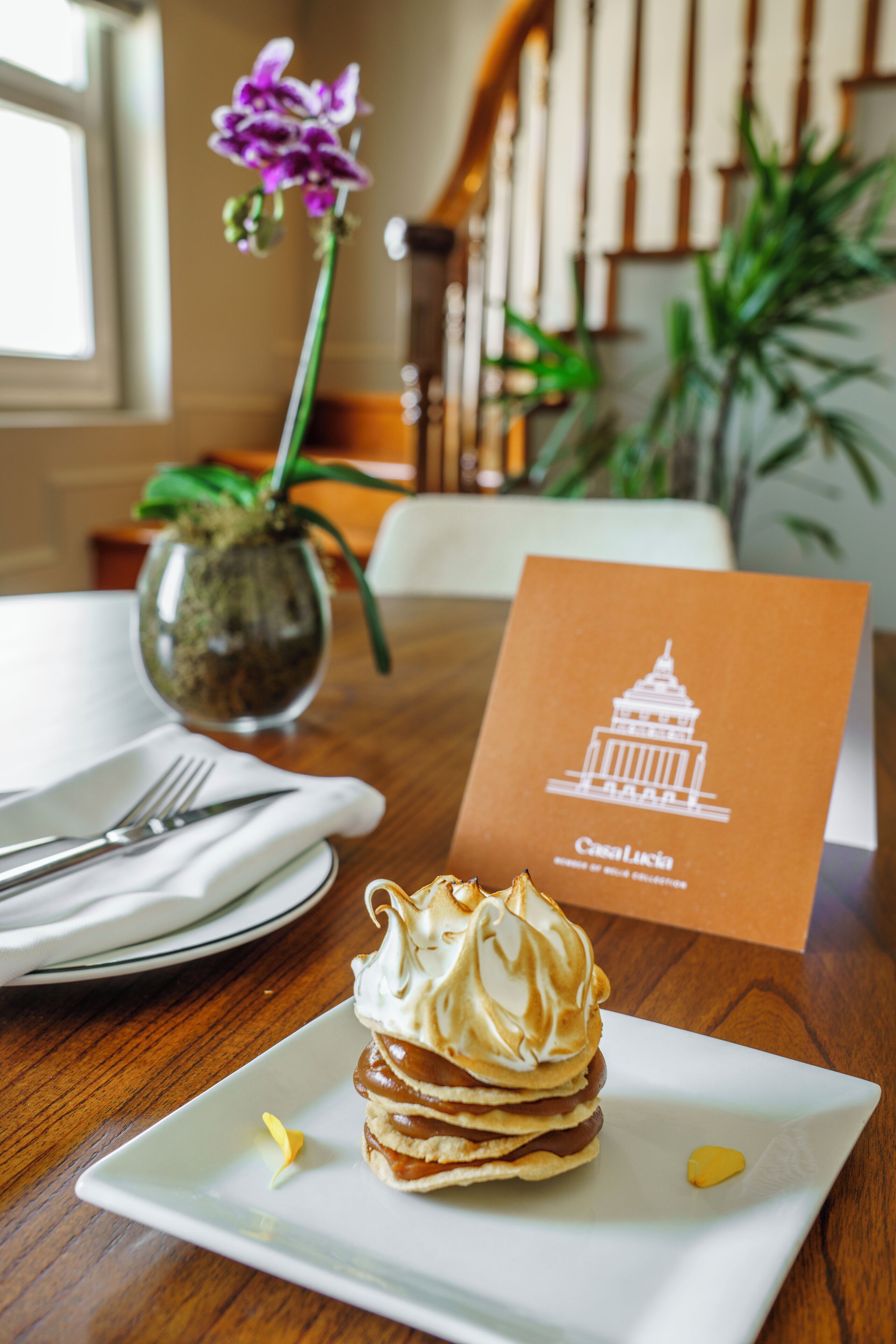 Un postre sobre un plato blanco, un jarrón con una flor púrpura y una tarjeta marrón con una ilustración de un edificio.