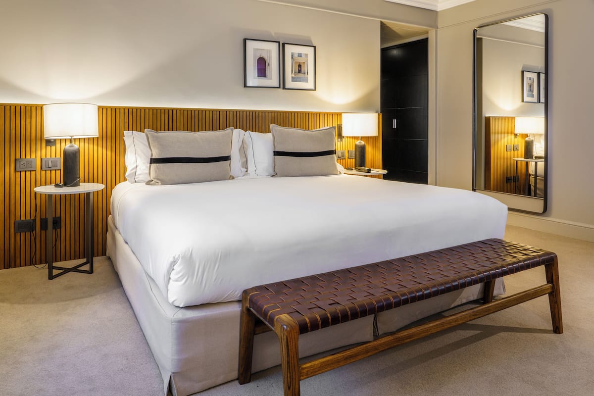 Una habitación de hotel con una cama grande, dos mesitas de noche con lámparas, un banco y un espejo grande.