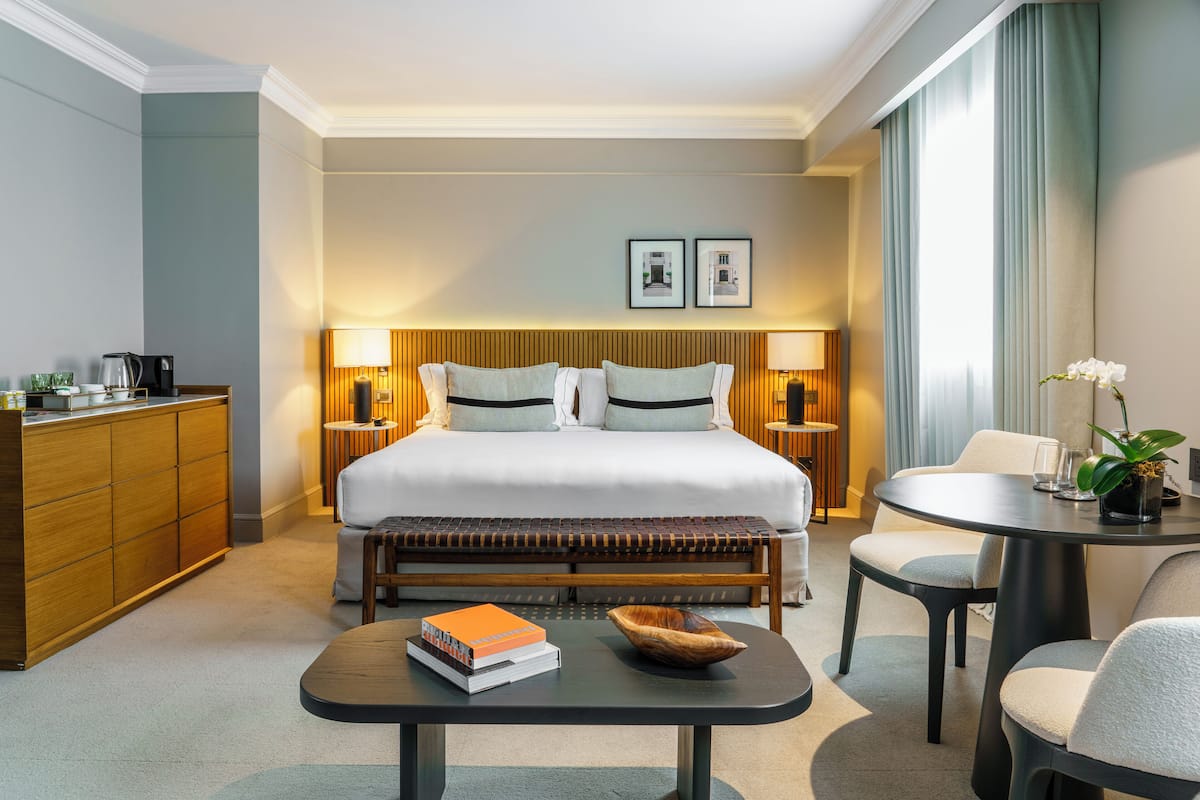 Habitación de hotel con cama, lámparas de noche, una mesita con libros, un cuenco y una silla.