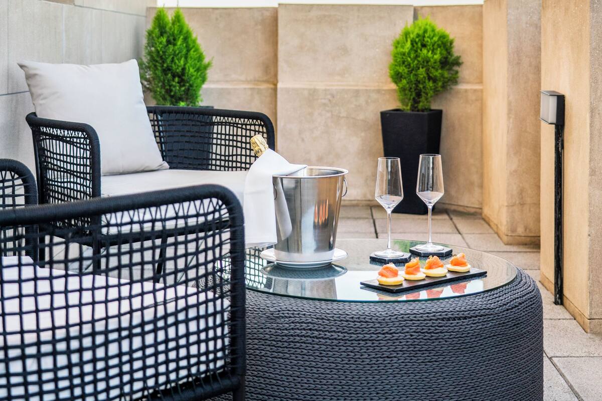 Una terraza en la azotea con mobiliario exterior, una mesa redonda con aperitivos y dos macetas.