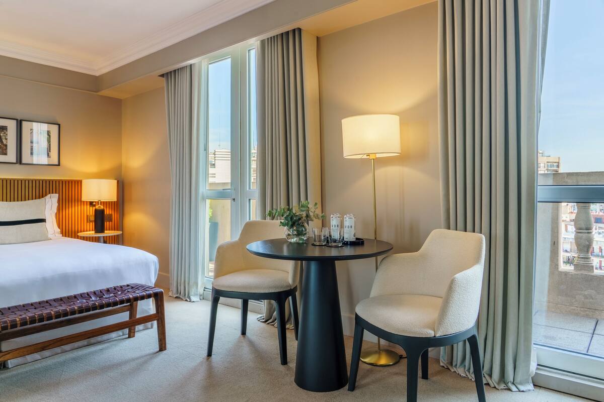 Una habitación de hotel con cama, un banco, dos sillas, una mesa y una lámpara.