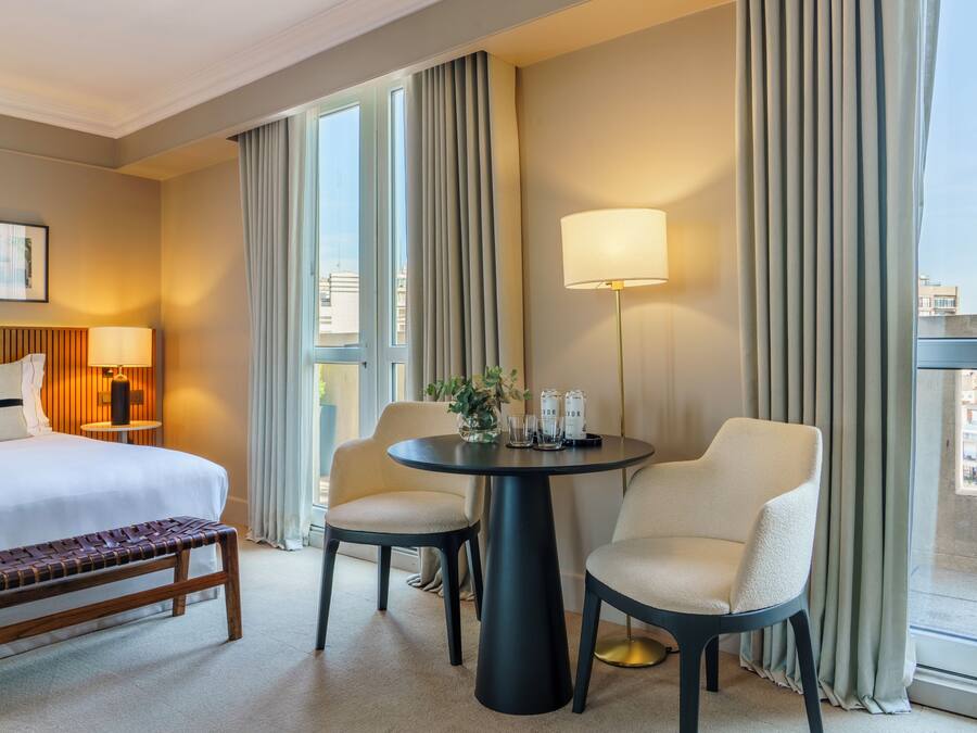 Una habitación de hotel con una cama, dos sillones, una mesita, una lámpara y una ventana con cortinas.