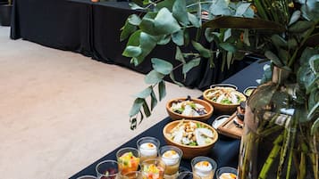 A table with various food and drink items, surrounded by large potted plants and a decorative glass vase.