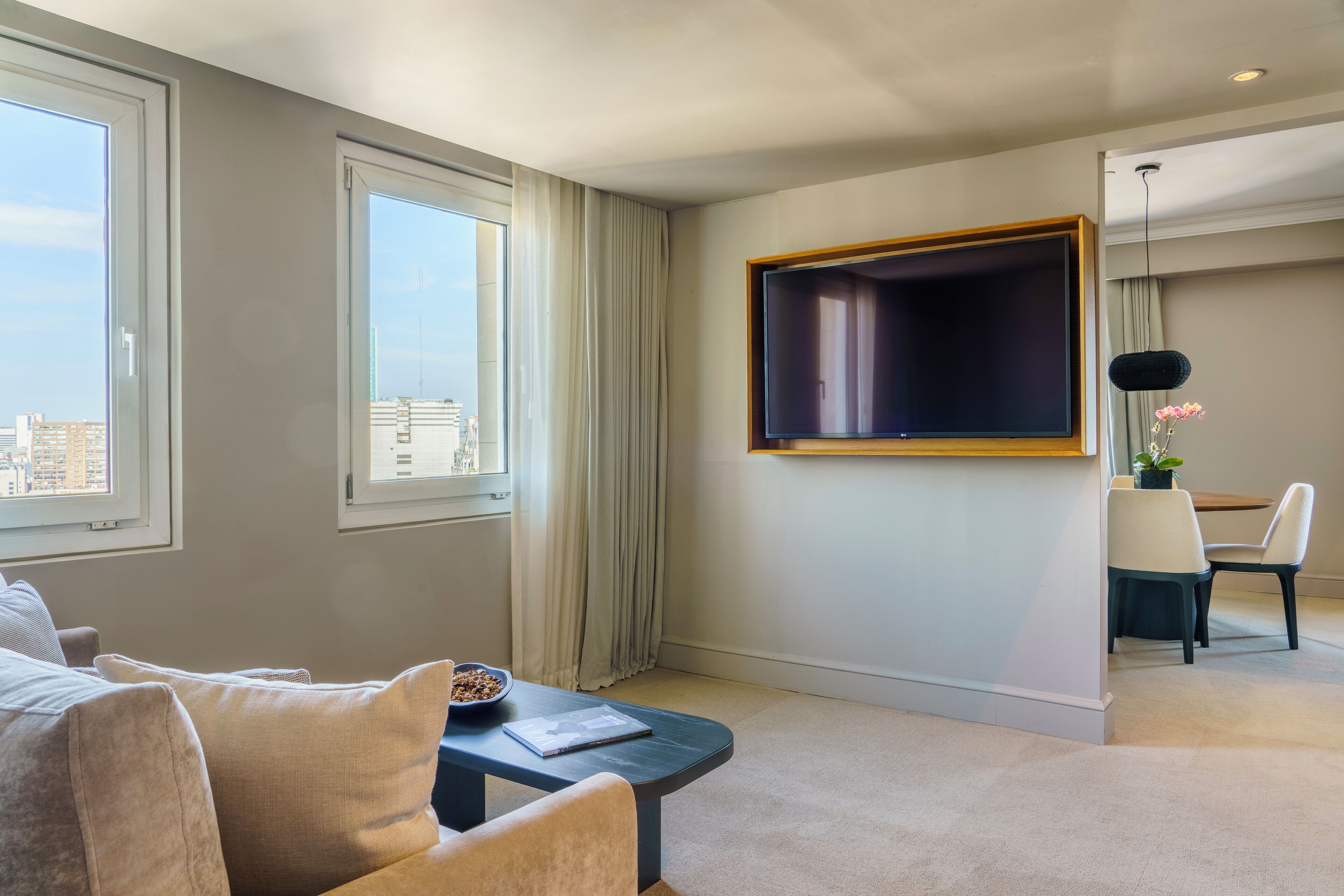 Quarto de hotel com uma televisão de tela grande na parede, um sofá, uma pequena mesa com tigela de cereal e uma área de refeições com mesa e cadeiras.