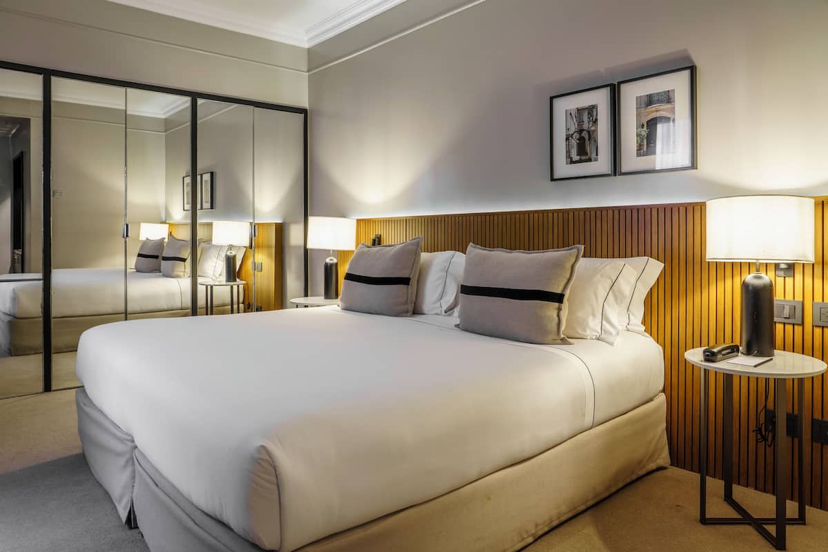 Un cuarto de hotel con una cama grande, un espejo, un cabecero de madera, dos mesitas de noche con lámparas y cuadros enmarcados en la pared.