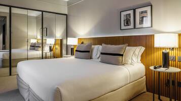 Un cuarto de hotel con una cama grande, un espejo, un cabecero de madera, dos mesitas de noche con lámparas y cuadros enmarcados en la pared.