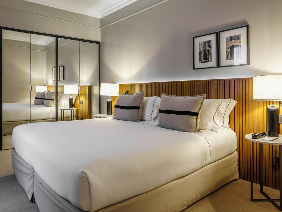 Un cuarto de hotel con una cama grande, un espejo, un cabecero de madera, dos mesitas de noche con lámparas y cuadros enmarcados en la pared.