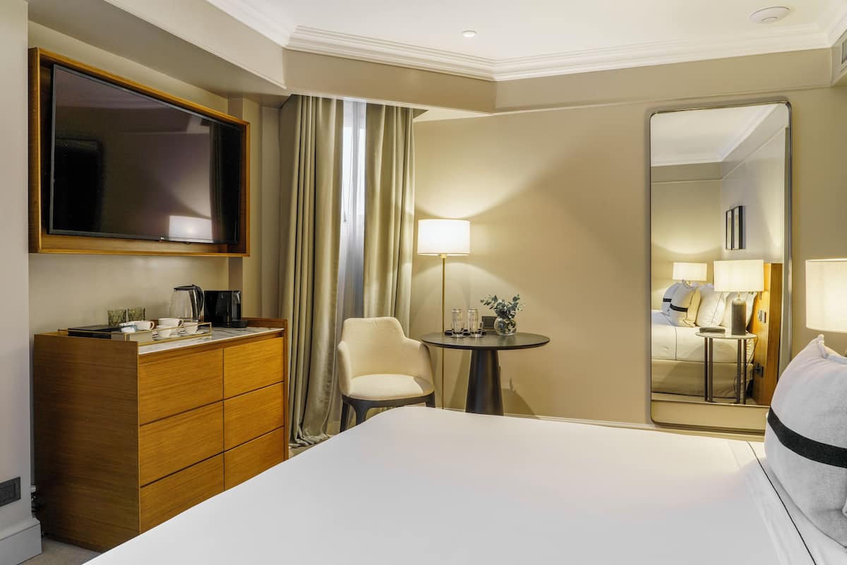 Habitación de hotel con cama, cómoda, televisor, silla, mesa con lámpara y espejo.