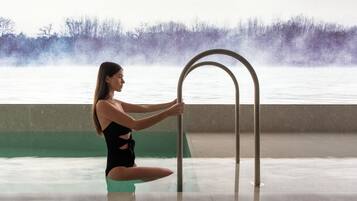 Una persona con traje de baño negro sentada junto a una piscina con una escalera metálica, con vistas a árboles y agua al fondo.