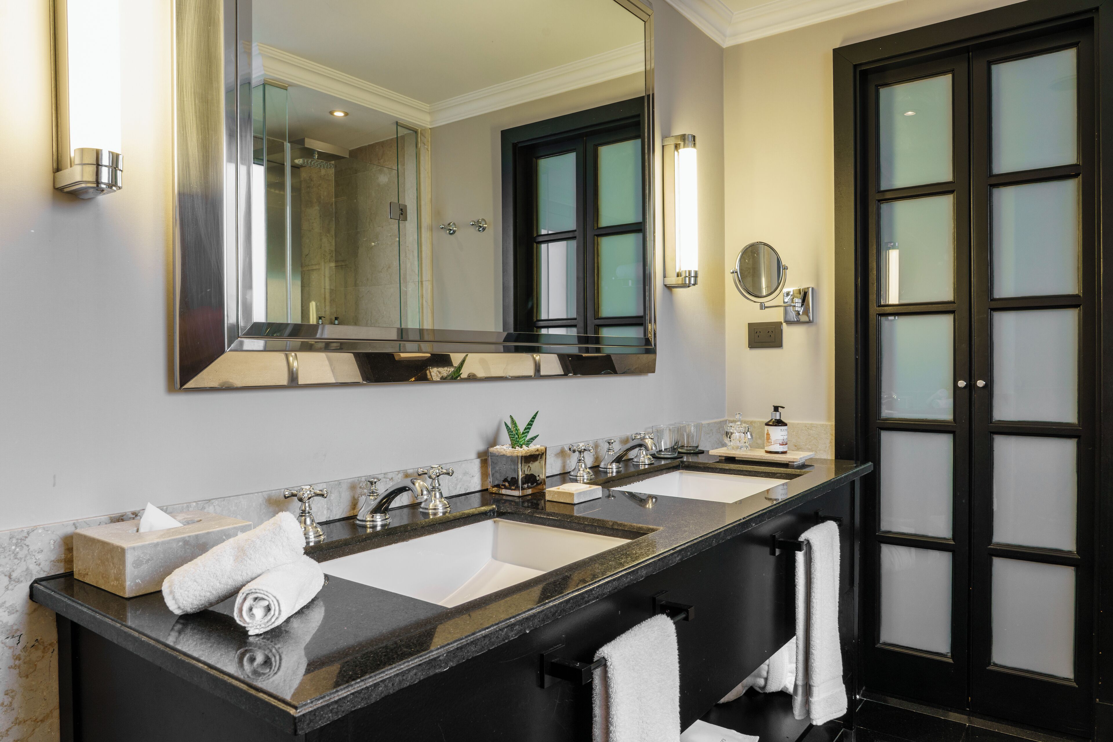 A modern bathroom with two sinks, a large mirror, and a black door with frosted glass panels.