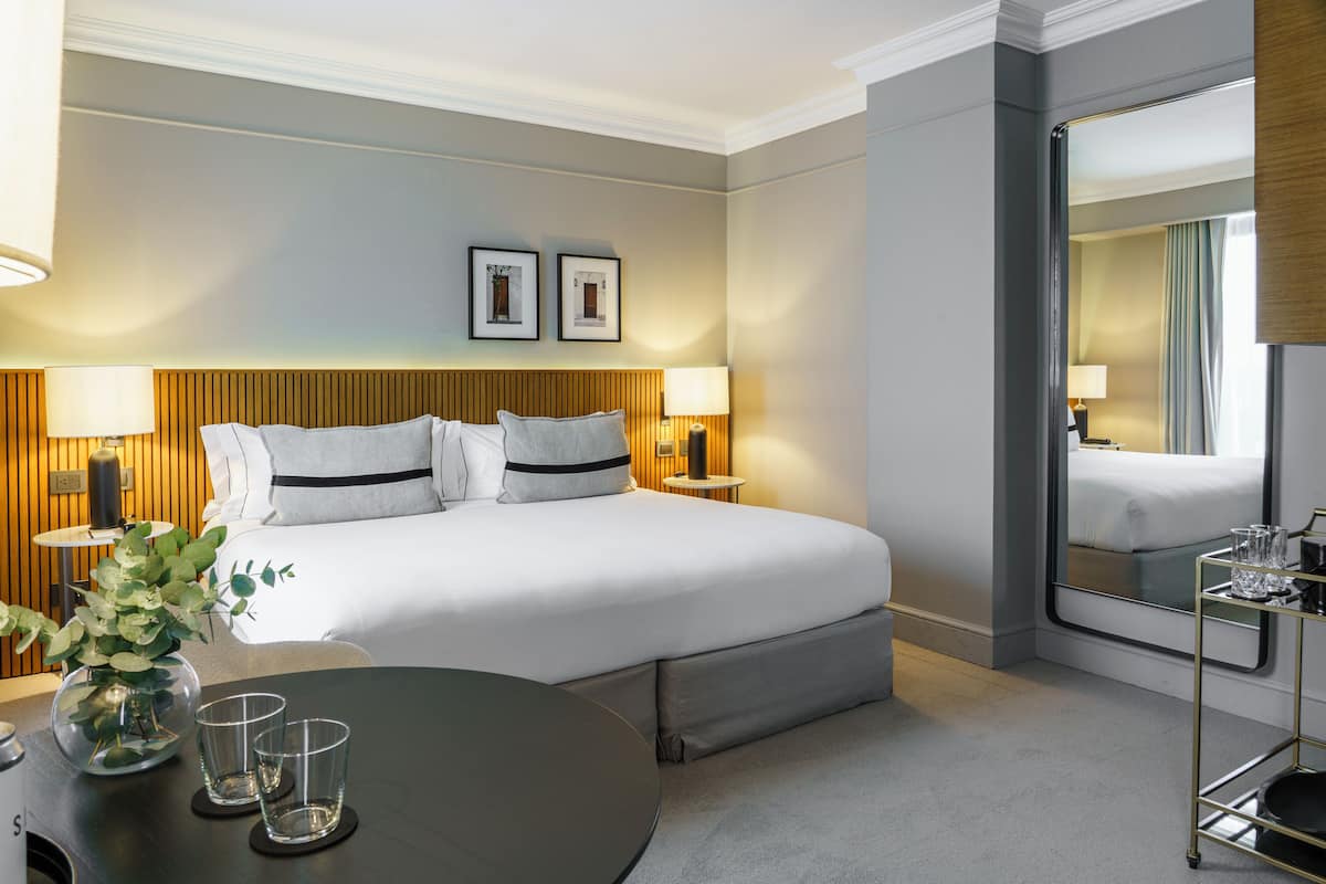 Habitación de hotel con una cama grande, dos mesitas de noche con lámparas, una mesa redonda con un florero y vasos, y un espejo en la pared.