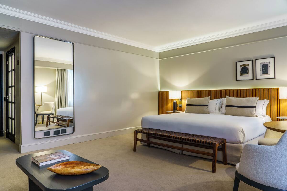 Habitación de hotel con una cama grande, un banco, una mesita de noche con un libro, un espejo y dos cuadros en la pared.
