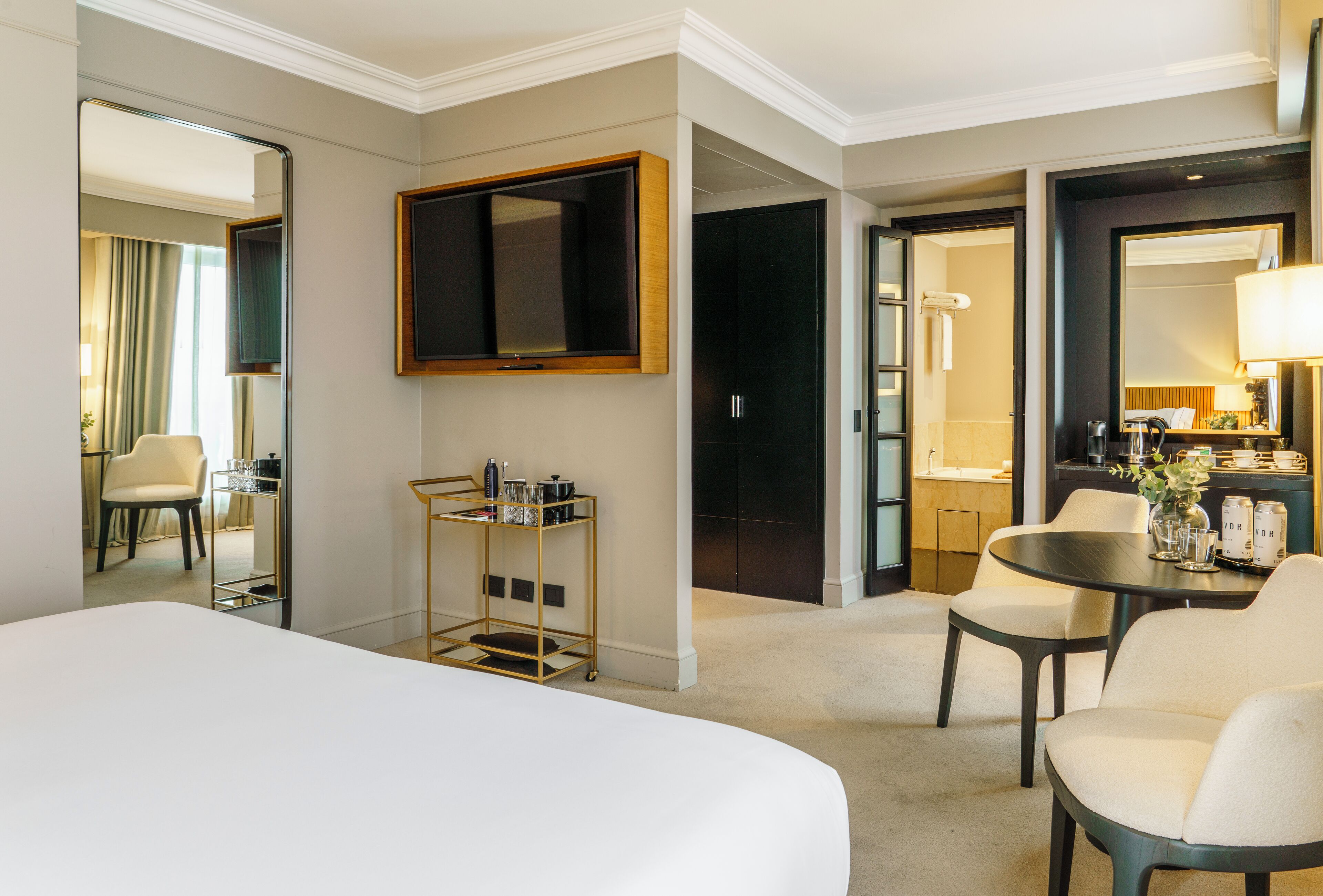 A hotel room with a large bed, a sitting area with a table and chairs, a small kitchenette, and a bathroom visible through an open door.