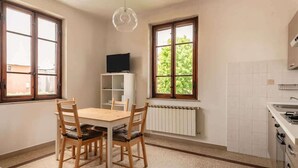A kitchen with a dining table and chairs, a TV on a shelf, and large windows with a view of greenery.