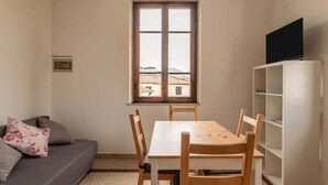 A small, well-lit room with a wooden table and chairs, a sofa with cushions, a television on a cabinet, and a window with a view of buildings.