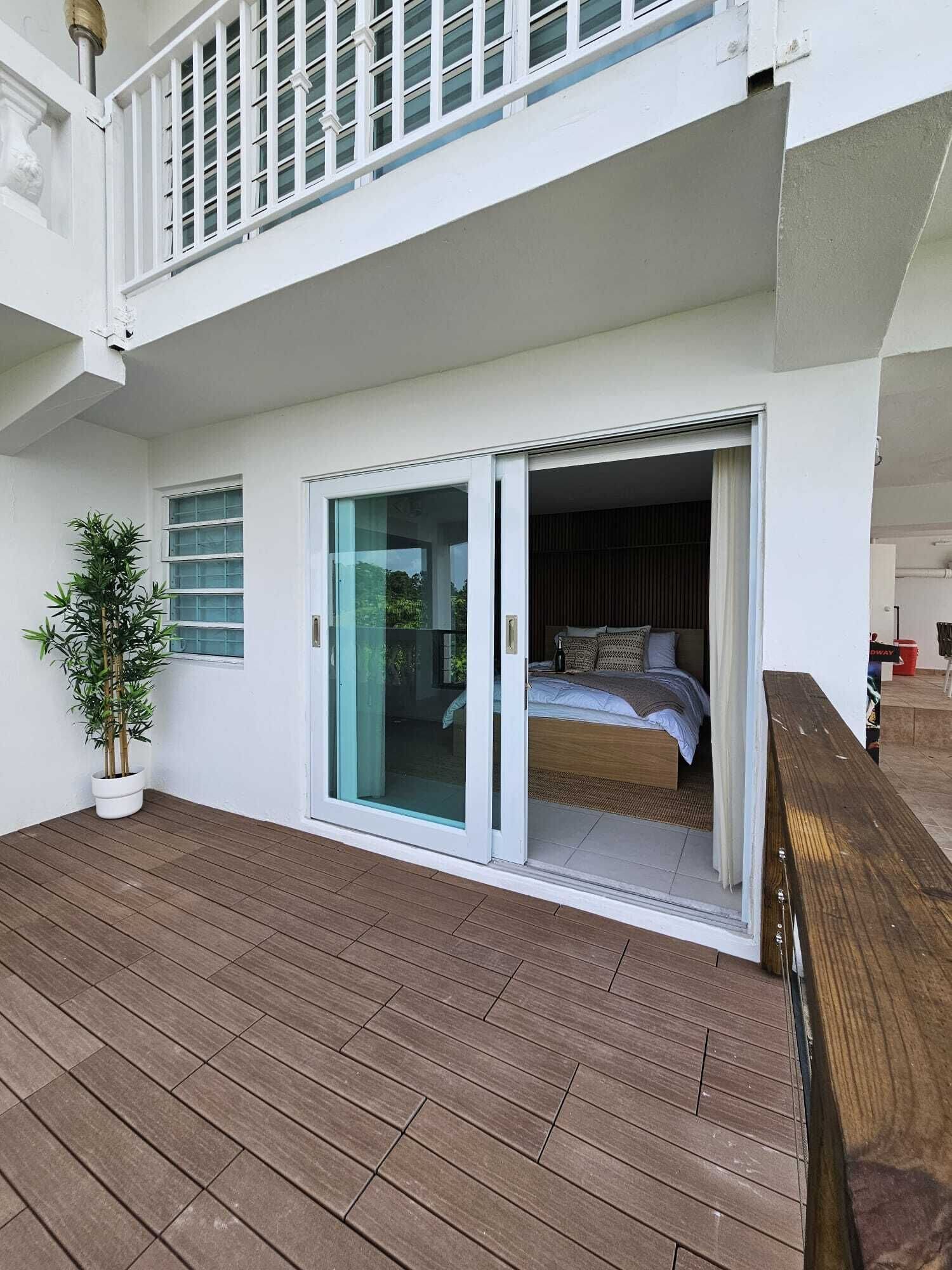 Een modern balkon met houten vloer, een bloempot en een slaapkamer zichtbaar door de glazen deur, met bed en gordijnen.