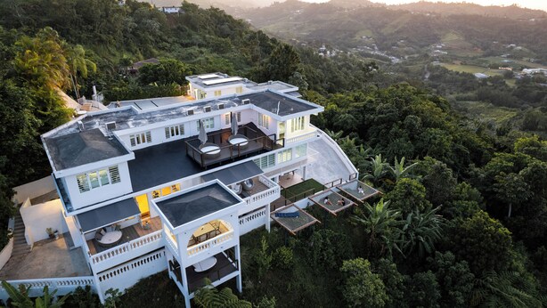 Ein mehrstöckiges Haus mit einer großen Terrasse und Sitzgelegenheiten im Freien, umgeben von üppiger Vegetation und Hügeln.