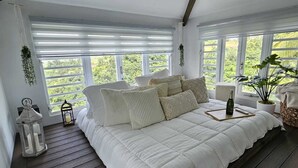 Ein Schlafzimmer mit einem großen Bett, Holzboden und mehreren Fenstern mit Blick auf Grünflächen.