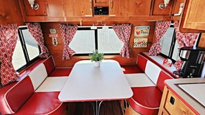 Interior compacto de uma motorhome, com área de refeições, assentos vermelhos e brancos, uma pequena mesa e uma cozinha equipada com micro-ondas.