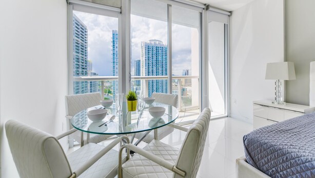 A modern hotel room with a balcony, a glass dining table, white furniture, and a bed with a patterned duvet. - Icon Tower 3 (Miami)