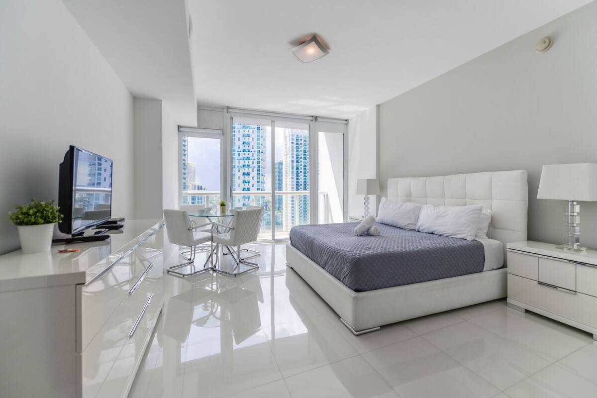 A modern hotel room with a large bed, a desk with a TV, and a balcony with a city view.
