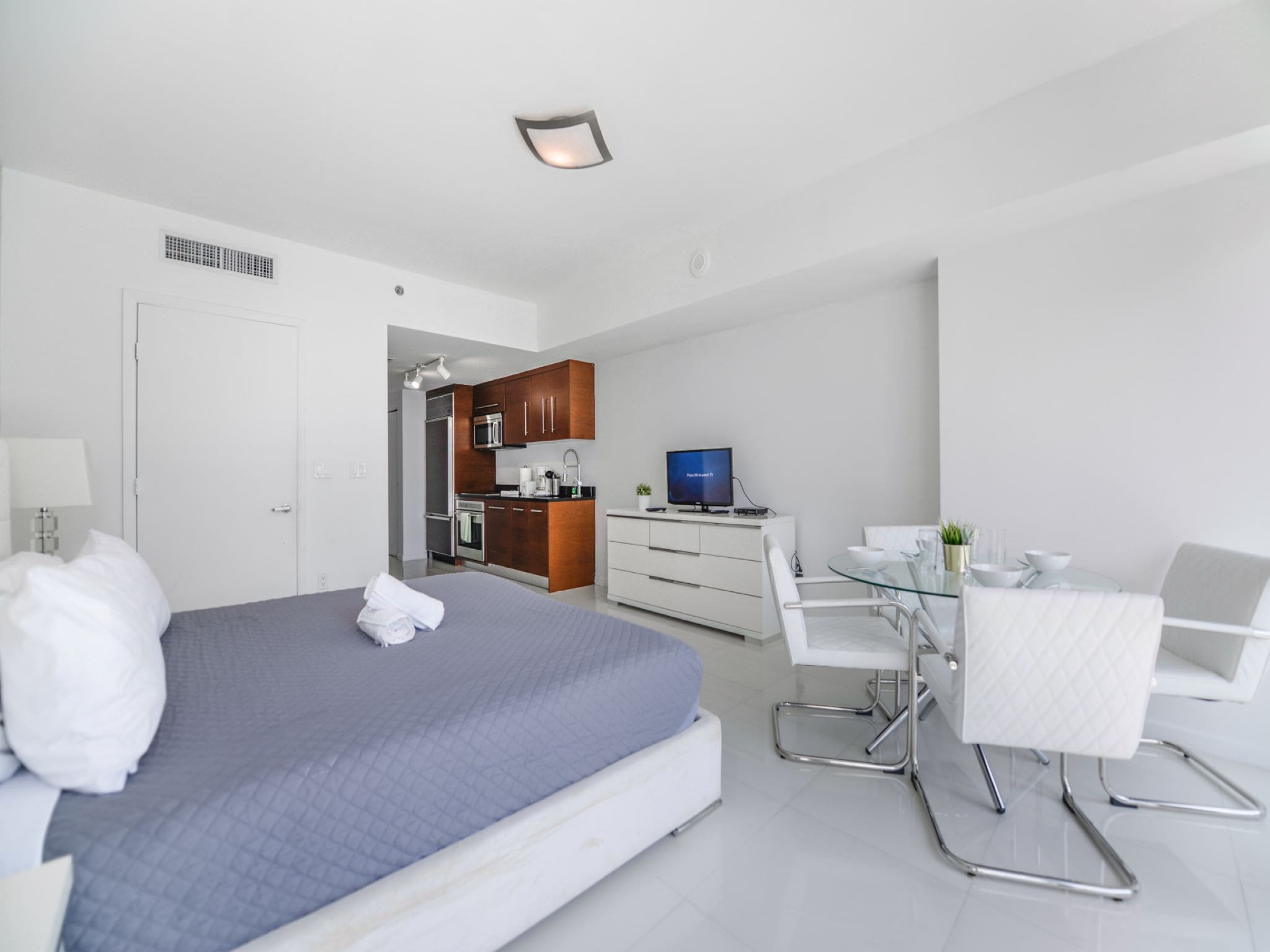 A modern hotel room with a bed, a small kitchenette, a dining area, and a TV.
