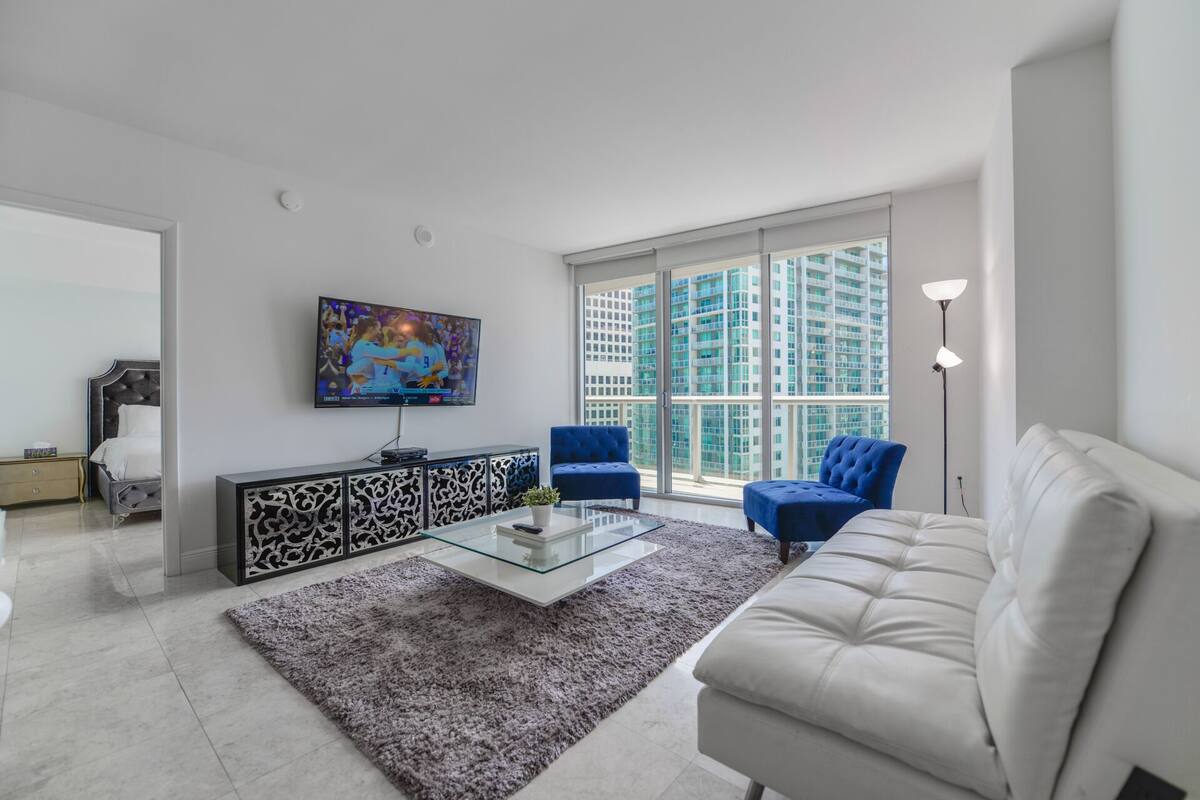 A modern living room with a glass coffee table, two blue armchairs, a white sofa, and a large window with a city view.