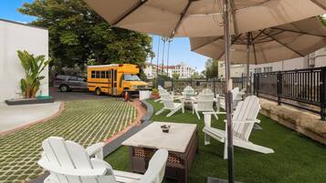 Uma área de pátio com cadeiras brancas, uma mesa e um ônibus ao fundo.
