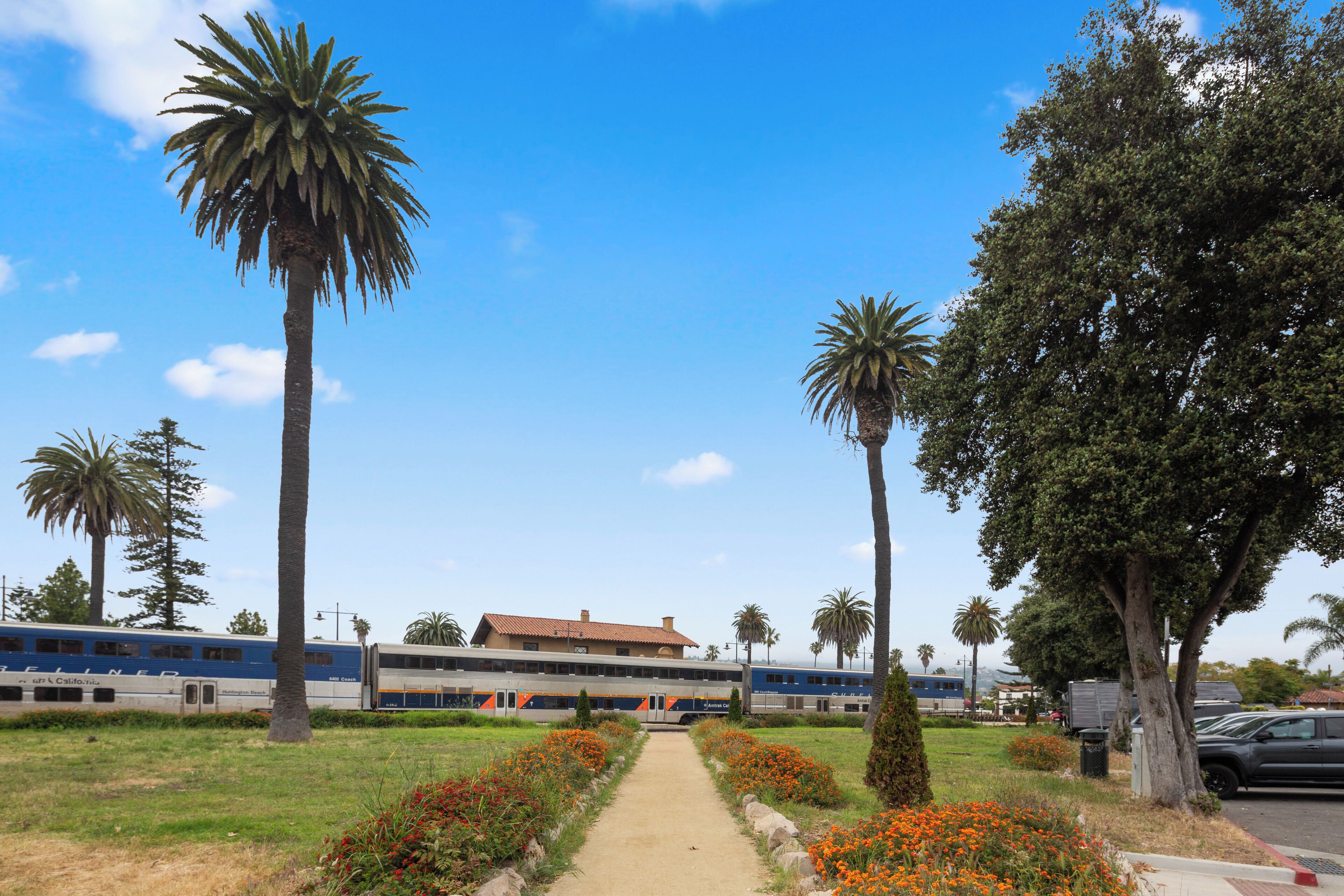 A train station with a clear pathway leading to a building, surrounded by palm trees and orange flowers.