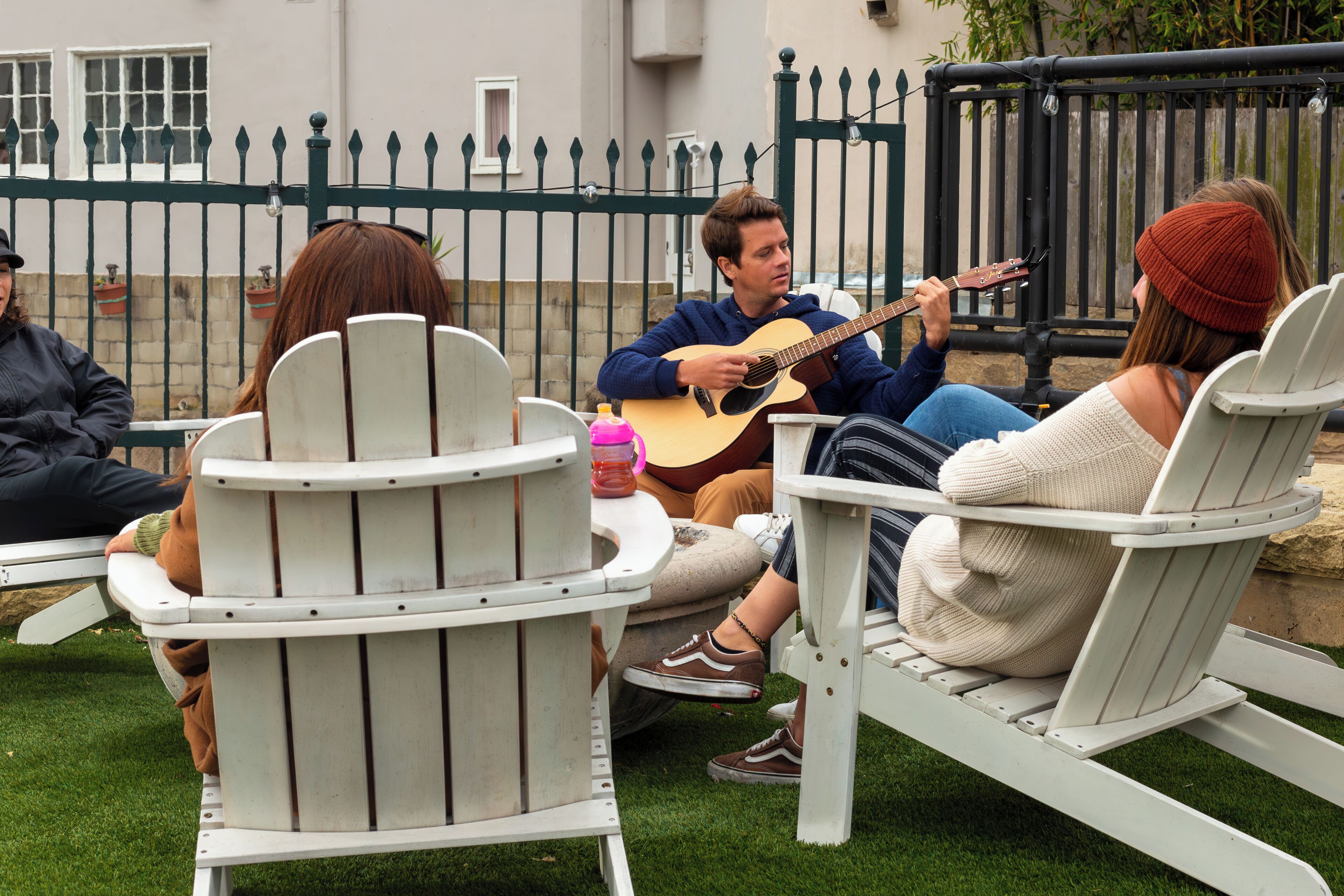 Un groupe de personnes est assis sur des fauteuils Adirondack blancs, à l’extérieur, et un homme joue de la guitare acoustique.