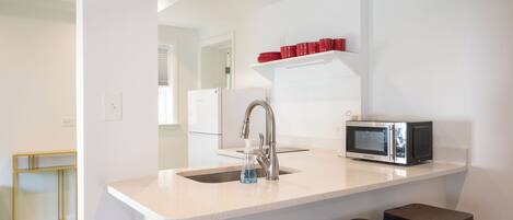 A kitchen with a white countertop, stainless steel sink, and a microwave. There are two stools and a shelf with red cups.
