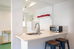 Una cocina con encimera blanca, pileta de acero inoxidable y microondas. Hay dos taburetes y una repisa con vasos rojos.