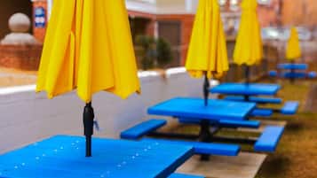 Une rangée de tables de pique-nique en plein air, surmontées de parasols jaunes, se trouve devant un bâtiment à la façade blanche.