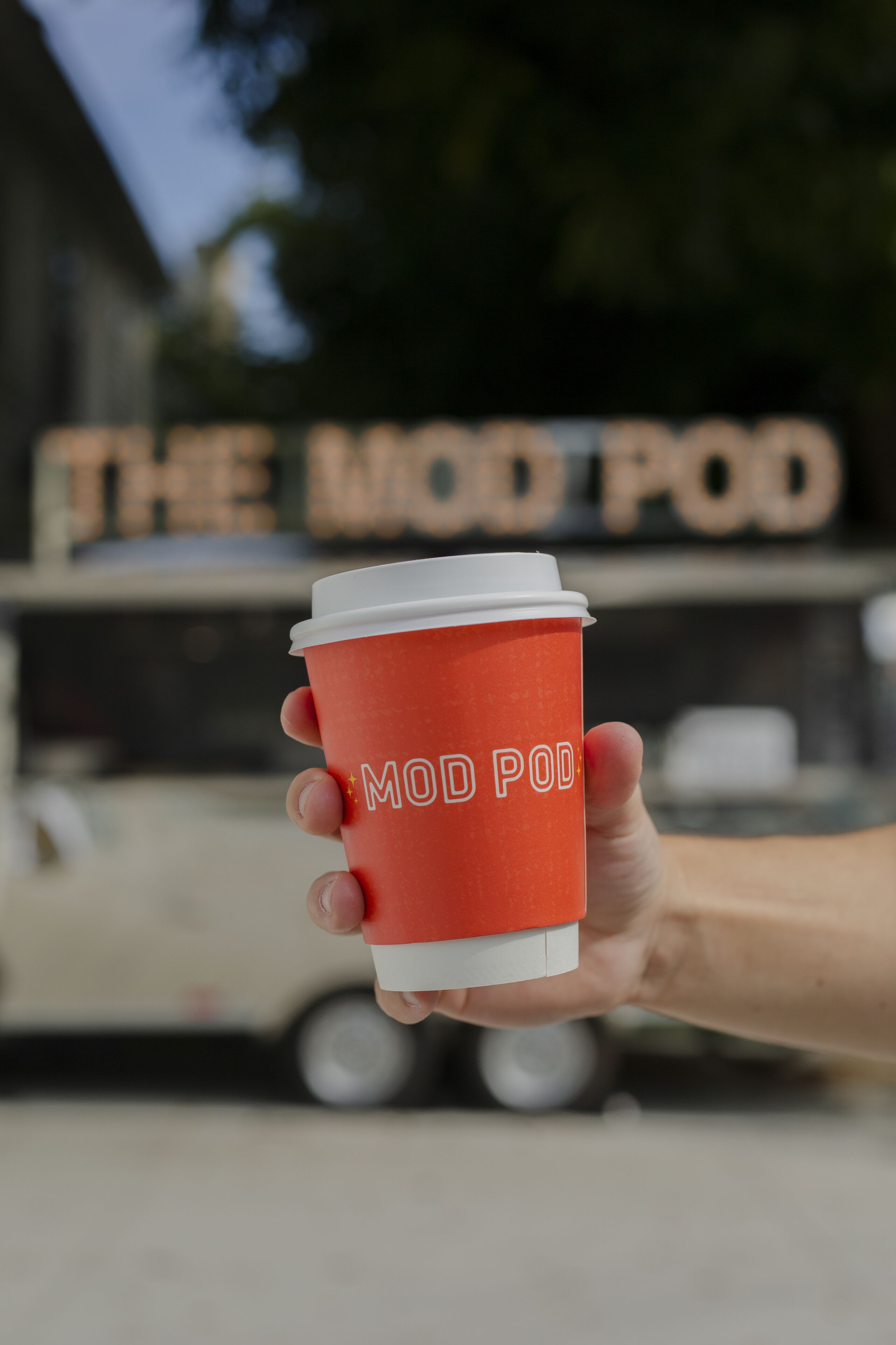 Eine Hand hält eine rote Kaffeetasse mit der Aufschrift "MOD POD", vor einem Imbisswagen mit der Beschilderung "THE WOO POD".